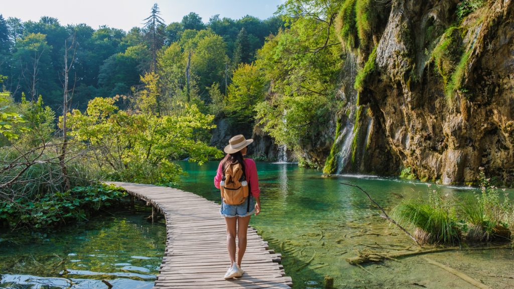 Eine Besucherin läuft auf einem Holzsteg an türkisfarbenem Wasser und kleinen Wasserfällen im Nationalpark Plitvicer Seen.