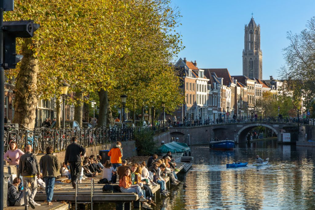 Menschen sitzen am Ufer der Oudegracht in Utrecht, im Hintergrund ragt der Domturm über die Häuser.