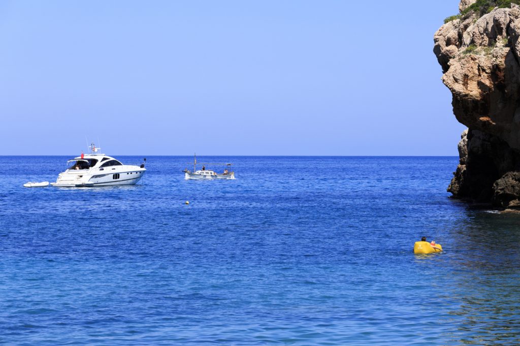 Yacht, Fischerboot und gelbes Tretboot liegen vor der Felsküste von Banyalbufar im ruhigen Mittelmeer.