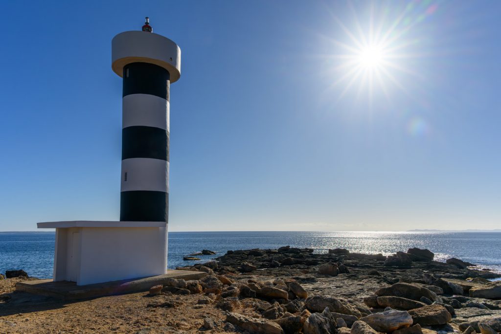 Der Far de s’Estalella ragt vor blauem Himmel auf, während die Sonne über dem Meer glitzert.