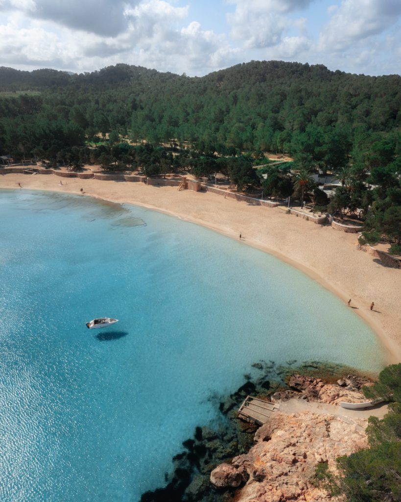 Blick auf die Cala Bassa mit hellem Sandstrand, flachem Wasser und einem Boot in der stillen Bucht.