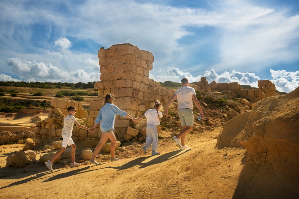 Familie wandert Hand in Hand durch eine sonnige Ruinenlandschaft auf Malta.