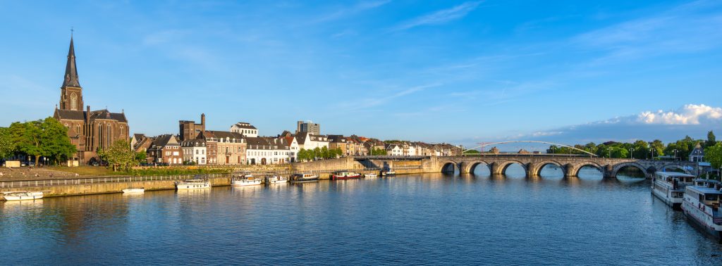 Die Maas fließt durch Maastricht, im Hintergrund stehen eine Kirche und eine Steinbrücke mit mehreren Bögen.