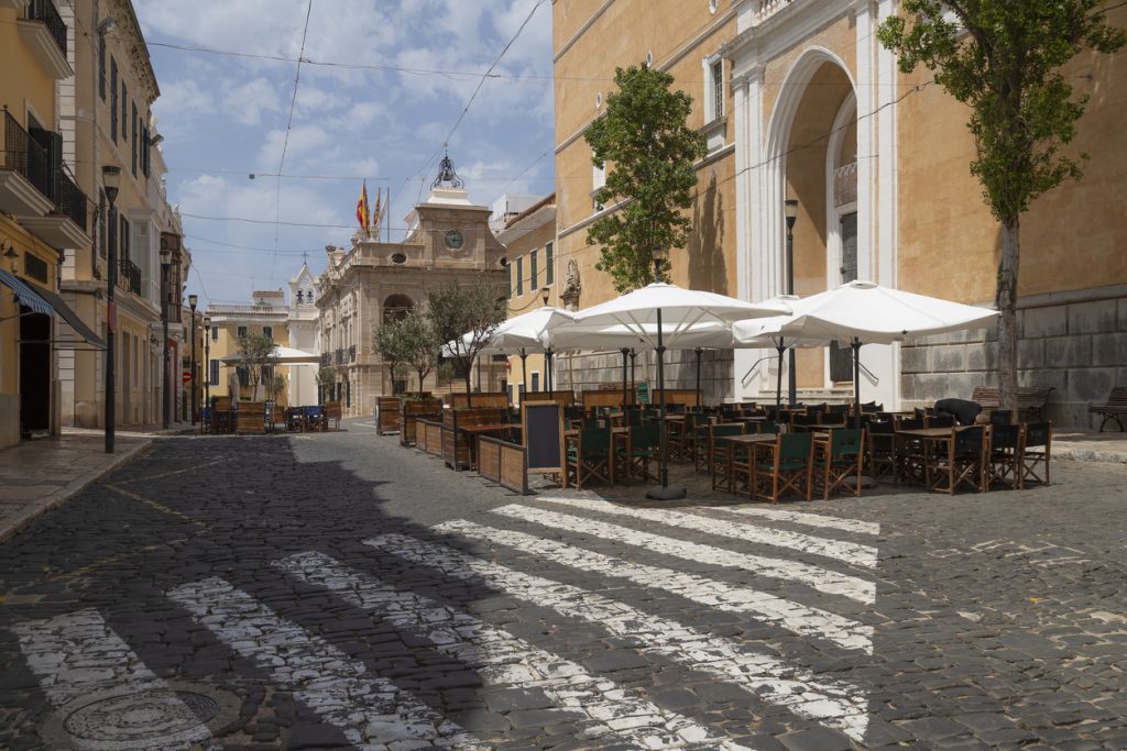 Leere Tische und weiße Sonnenschirme auf einem Platz in Maó auf Menorca.