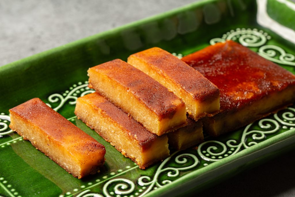 Rechteckige, goldbraun gebackene Stücke Turrón liegen ordentlich geschichtet auf einer dekorativen grünen Keramikplatte.