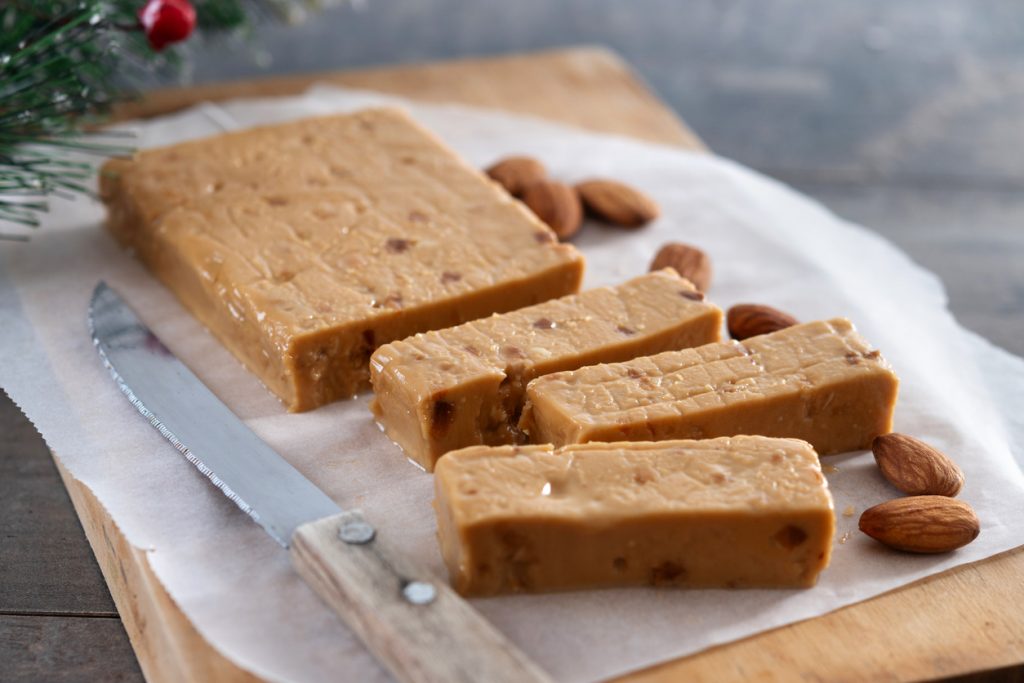 Weicher Turrón mit Mandeln liegt in Scheiben geschnitten auf einem Holzbrett neben einem Messer.