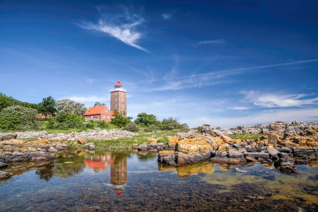 Alter Leuchtturm mit roten Dächern an der Felsküste von Bornholm und Spiegelung im Wasser.