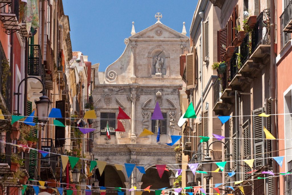 Enge Straße in Cagliari mit bunten Wimpeln und historischer Kirchenfassade.