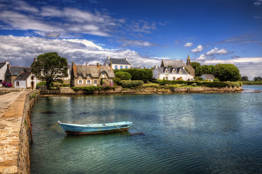 Kleine Steinhäuser mit Schieferdächern stehen an einem stillen Wasserarm in der Bretagne, vor dem ein blaues Boot liegt.