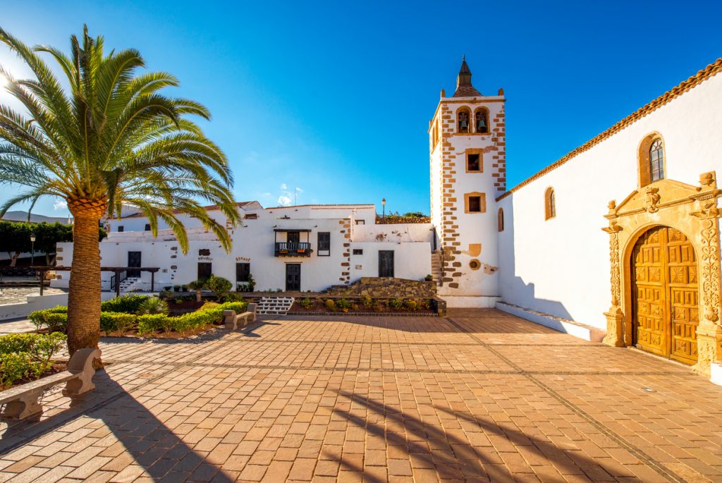 Sonniger Platz mit Palmbaum und weißer Kirche mit Glockenturm in Betancuria auf Fuerteventura.