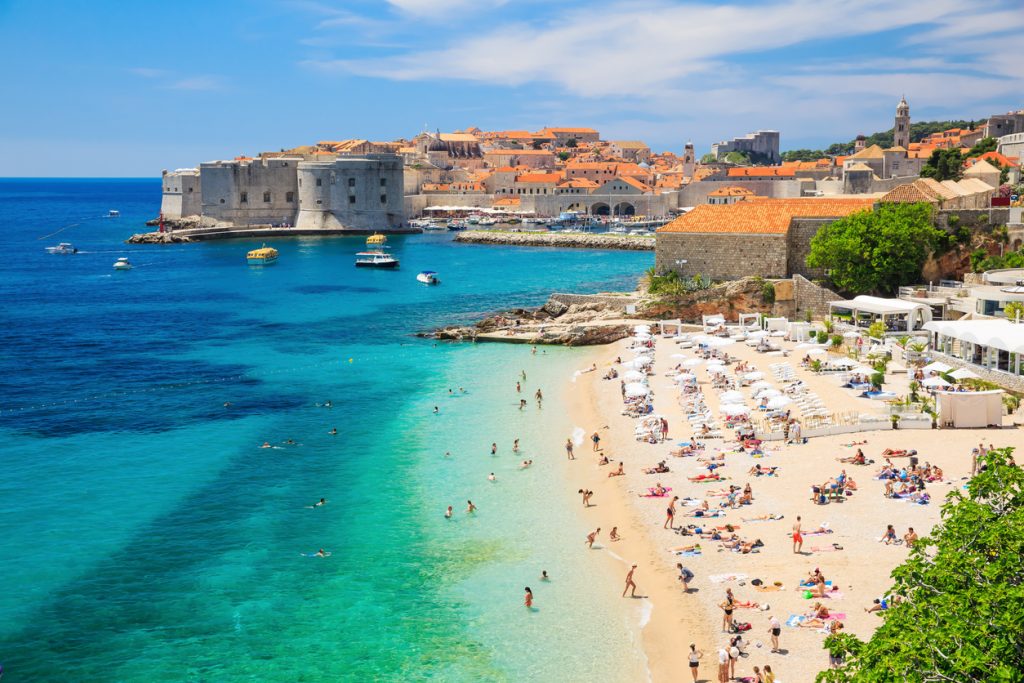Ein Strand in Dubrovnik liegt an kristallklarem Wasser mit Blick auf die Altstadt und Festungsanlagen.
