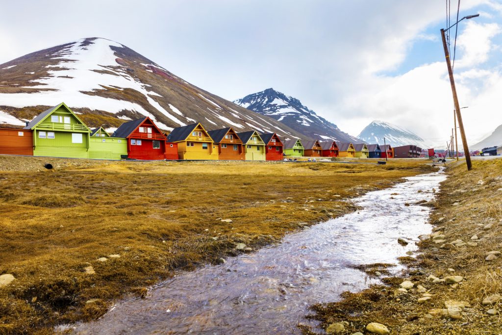 Eine lange Reihe bunter Häuser steht an einem Hang mit Restschnee und Bachlauf auf Spitzbergen.