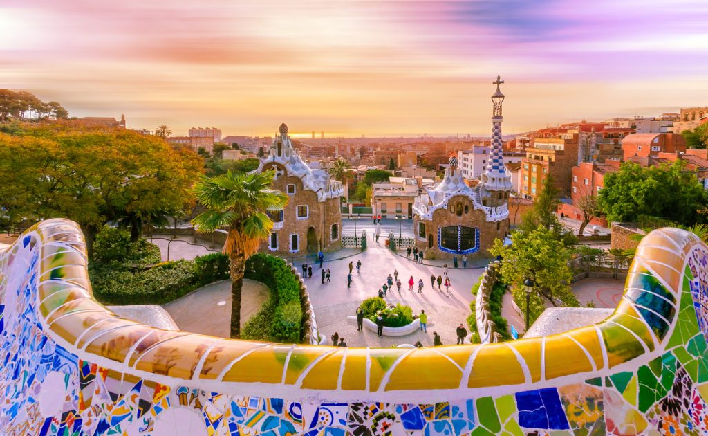 Blick vom Park Güell in Barcelona über die Stadt mit buntem Mosaikgeländer und Gaudí-Bauten.