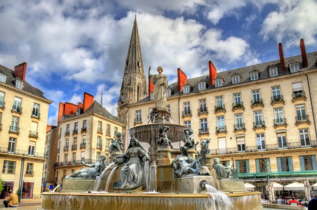 Der Brunnen auf der Place Royale in Nantes sprudelt vor klassischen Figuren, dahinter ragt die Kirche Saint-Nicolas.