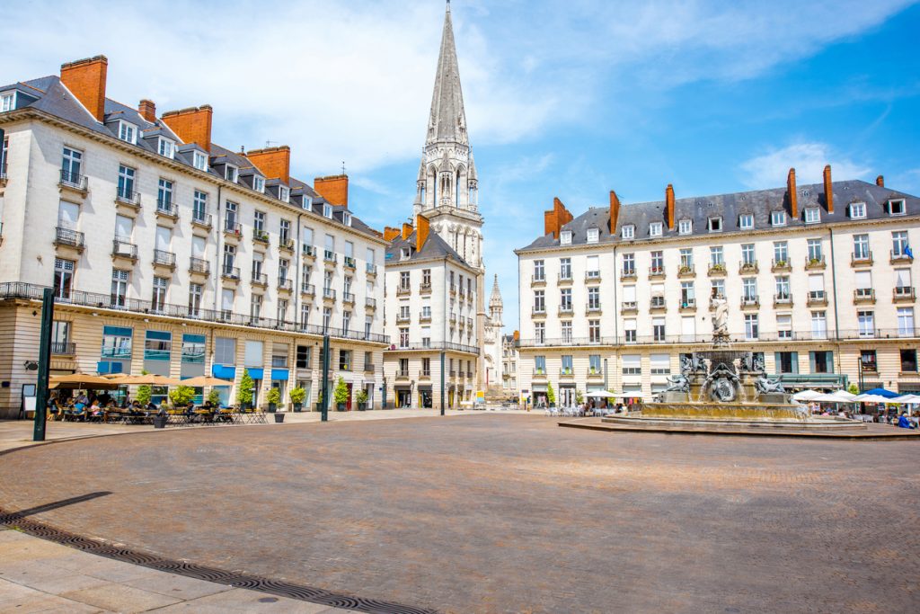 Die Place Royale in Nantes zeigt offene Platzfläche, Cafés und den Brunnen mit der Kirche Saint-Nicolas im Hintergrund.