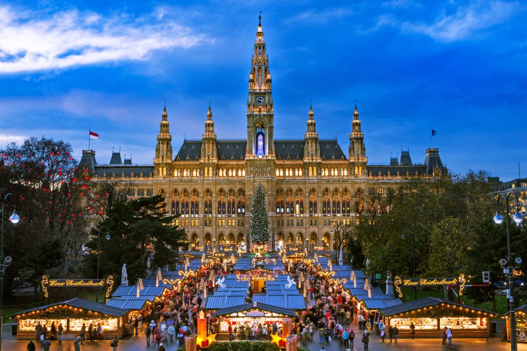 Weitläufiger Weihnachtsmarkt vor dem erleuchteten Wiener Rathaus in der Dämmerung.