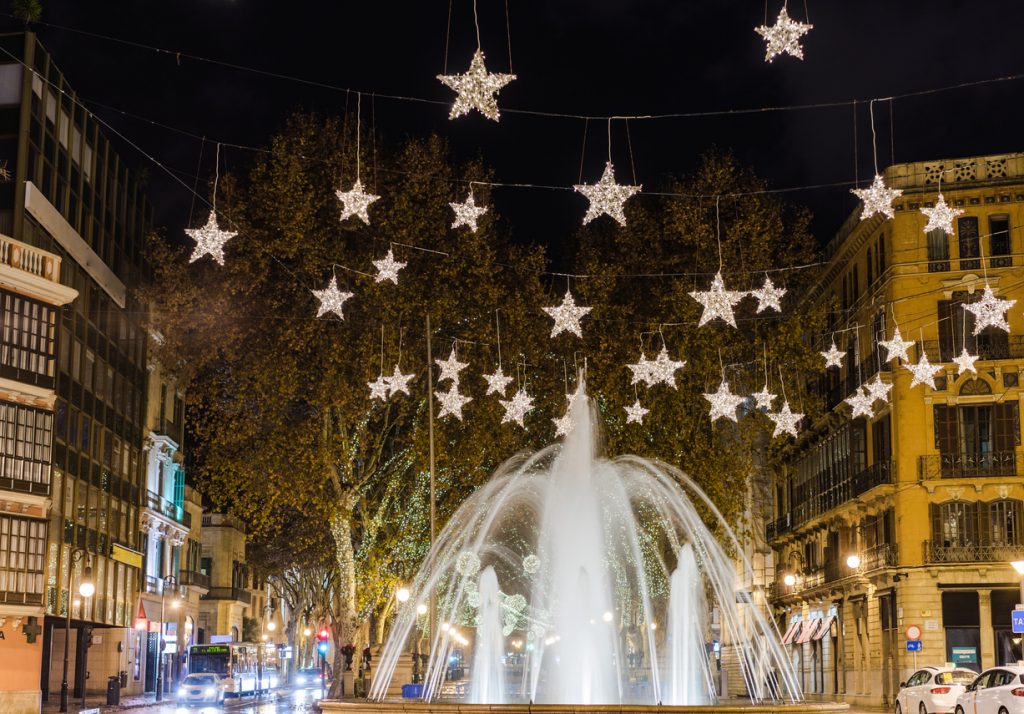 Weißer Springbrunnen in einer Stadt auf Mallorca, über dem zahlreiche leuchtende Stern-Dekorationen eines Weihnachtsmarktes hängen.