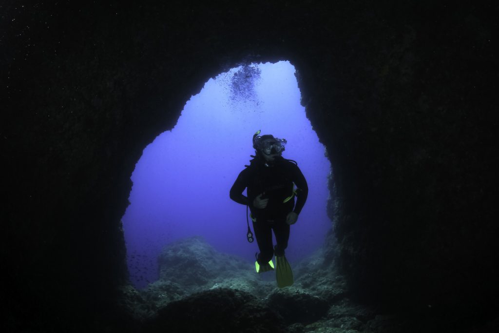Ein Taucher schwimmt durch einen Felsbogen und blickt ins helle Wasser beim Tauchen auf Mallorca.