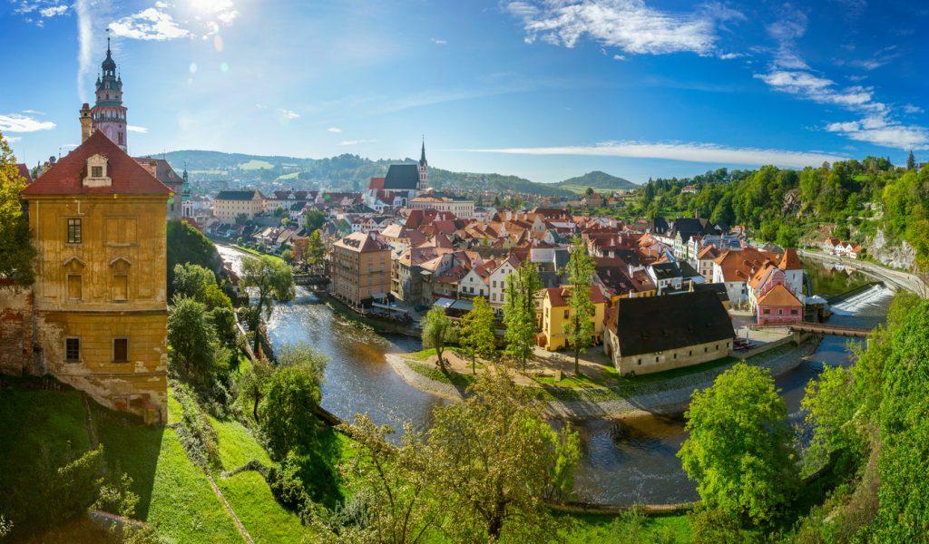 Blick über Český Krumlov mit Flussschleife, roten Dächern und Turm unter blauem Himmel.