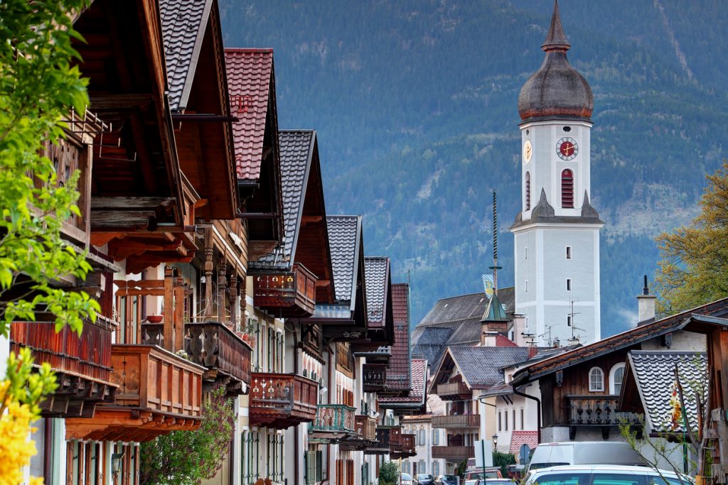 Holzfassaden und Balkone führen auf einen weißen Kirchturm vor Bergkulisse in Garmisch-Partenkirchen zu.