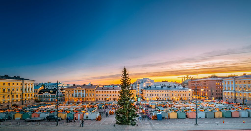 Viele Marktstände und ein großer Weihnachtsbaum stehen auf einem Platz in Helsinki bei Sonnenuntergang.