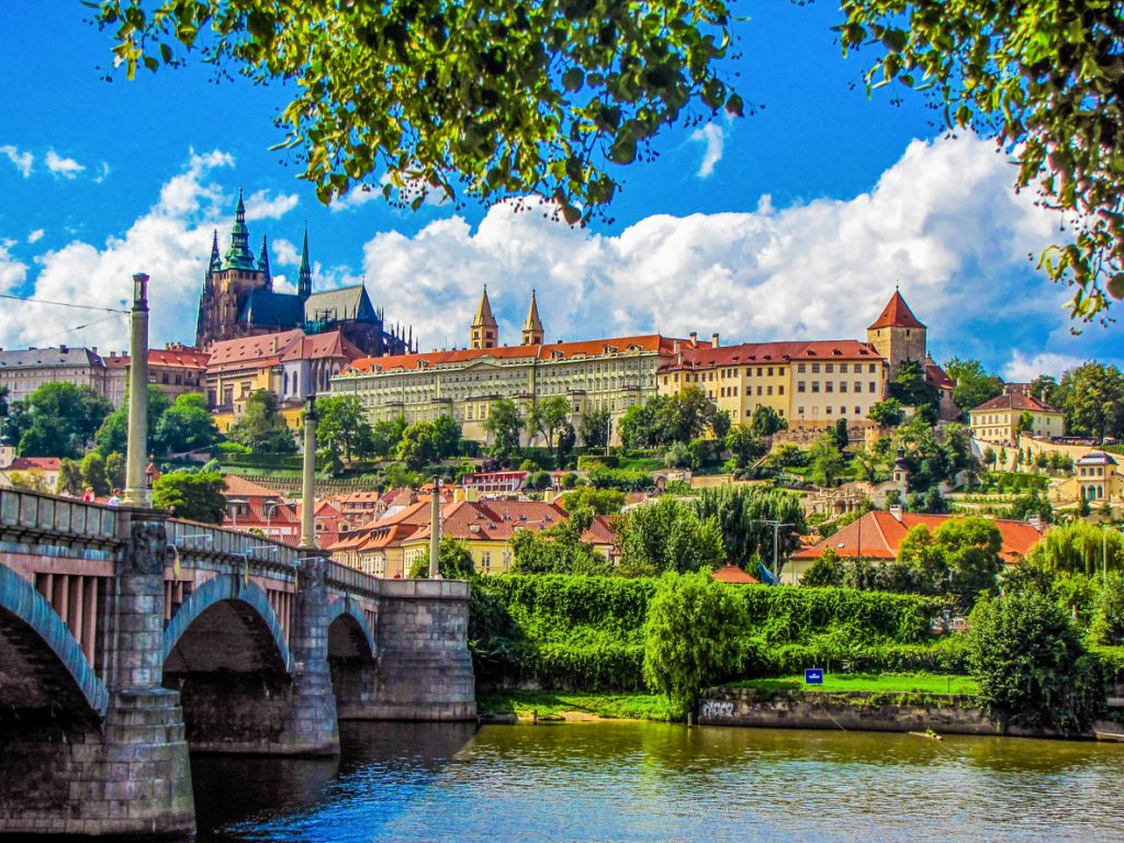 Blick auf die Prager Burg über den Fluss mit einer Steinbrücke im Vordergrund.