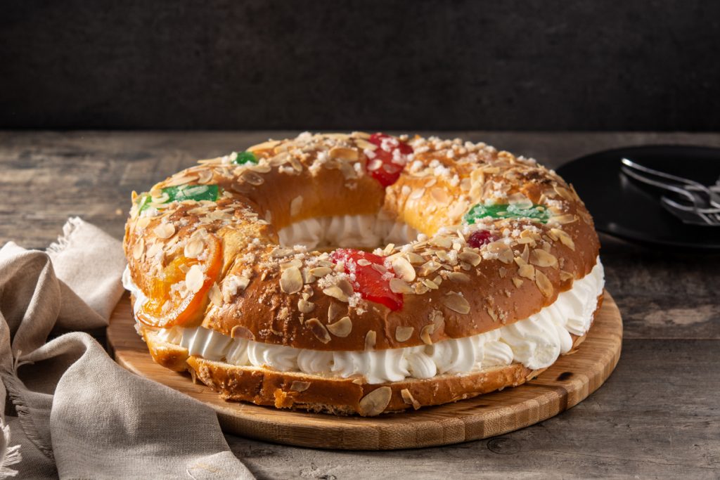Ein ganzer Roscón de Reyes mit Sahnefüllung ist mit kandierten Früchten und Mandeln dekoriert und liegt auf einem Holzbrett.