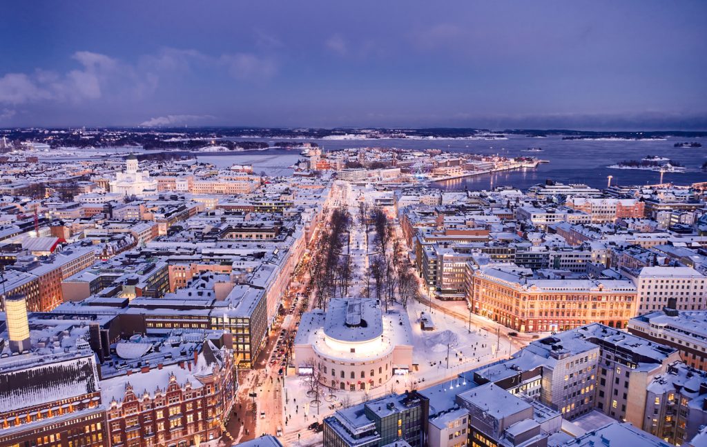 Ein verschneites Stadtpanorama von Helsinki zeigt breite Straßen, warme Lichter und die Bucht im Hintergrund.