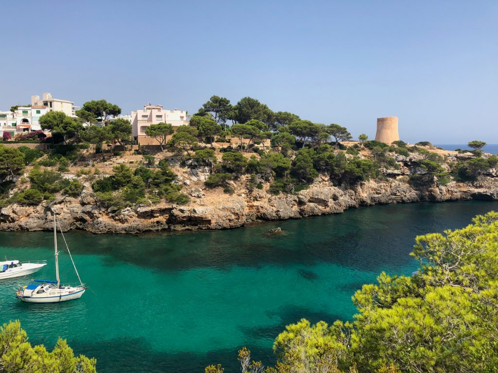 Segelboot in einer ruhigen Bucht, dahinter Küste mit Häusern und der Torre de Cala Pi.