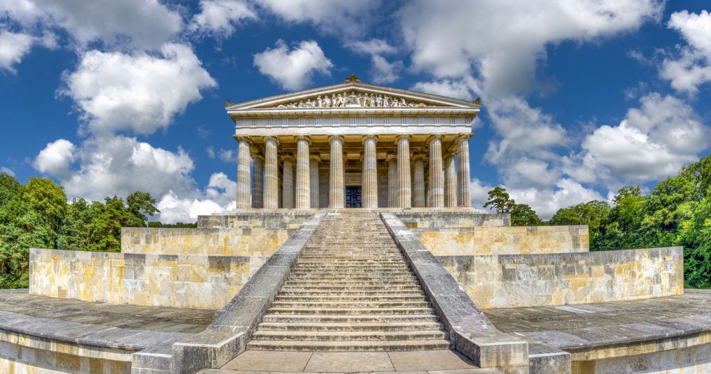 Die Walhalla bei Regensburg steht mit breiter Treppe und Säulen vor einem Wolkenhimmel.