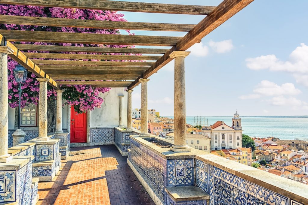 Veranda mit Azulejo-Kacheln, Holzpergola und pinker Bougainvillea mit Blick über Dächer und den Tejo in Lissabon.