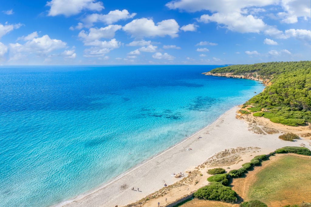 Der Strand Binigaus verläuft als helle Küstenlinie neben türkisblauem Wasser und grüner Macchia.