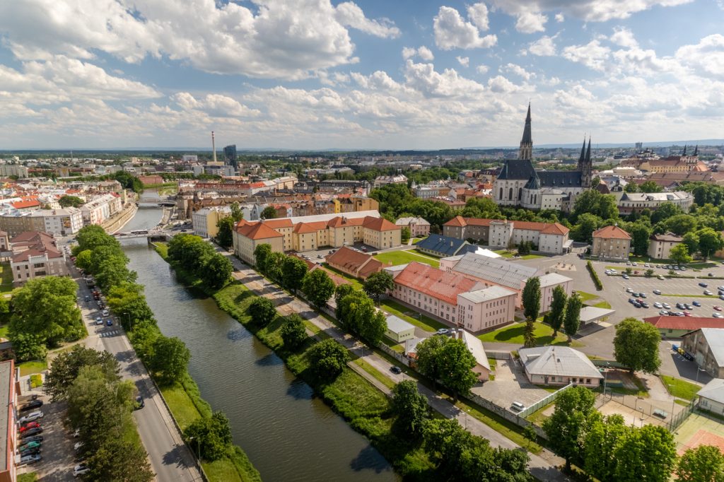 Olmütz mit Flusslauf, grünen Ufern und großen Kirchenbauten in der Ferne bei sonnigem Himmel.