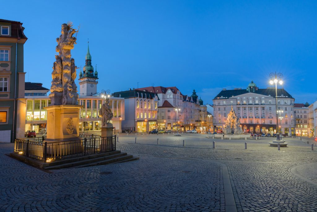 Zentraler Platz in Brünn zur blauen Stunde mit barocken Statuen, Laternen und historischen Gebäuden.