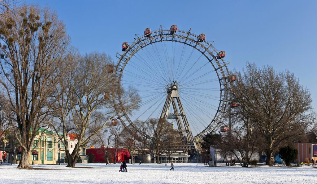 Das Wiener Riesenrad steht hinter kahlen Bäumen über einer verschneiten Wiese im Prater.