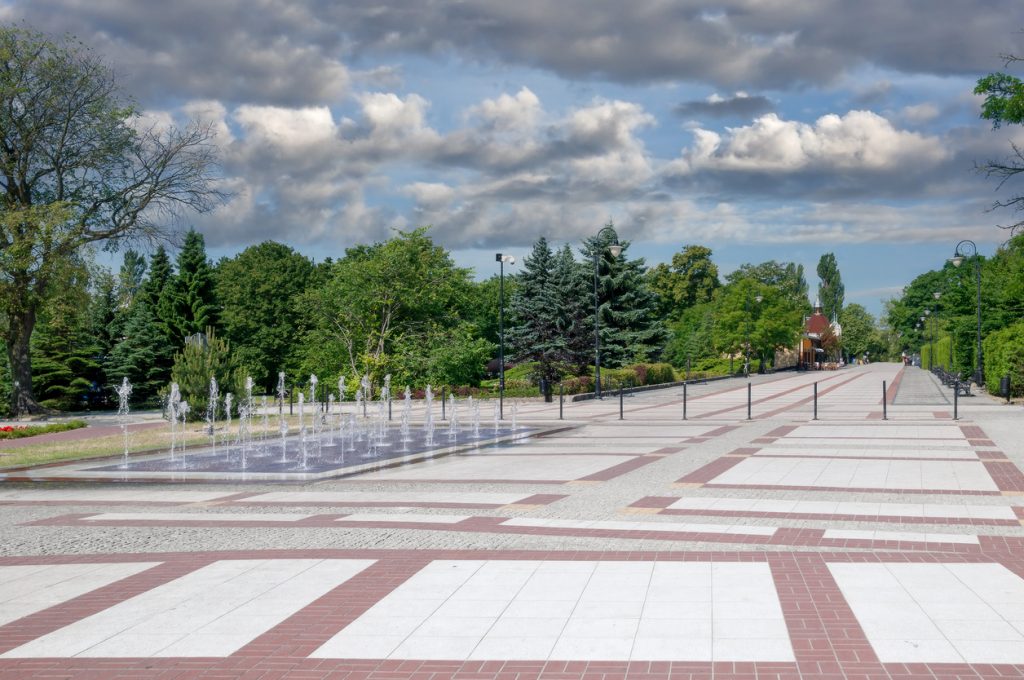 Eine gepflasterte Allee mit Fontänen und Bäumen führt geradeaus durch einen Parkbereich.