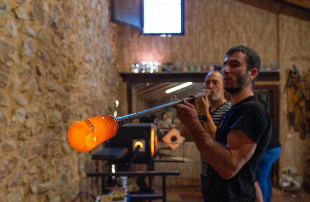 Ein Glasbläser bläst in einer Glasbläserei eine orange glühende Glasmasse an einer langen Pfeife.