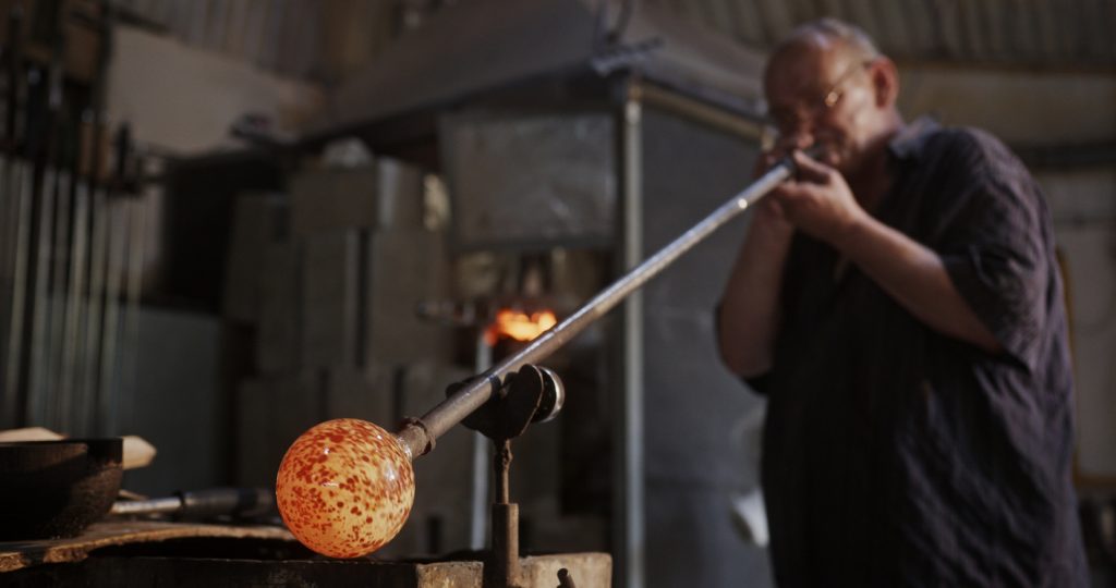 Ein Glasbläser arbeitet in einer Glasbläserei mit einer Pfeife, an deren Ende eine gesprenkelte Glasblase sitzt.