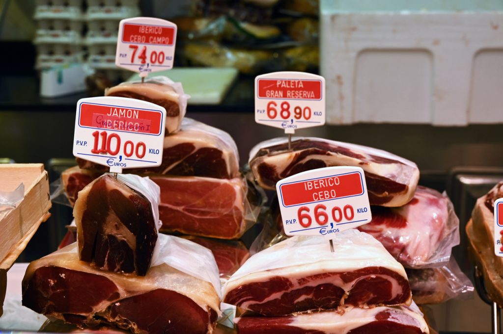 Stücke von Jamón und iberischem Schinken mit Preisschildern an einer Theke im Mercat de l’Olivar.