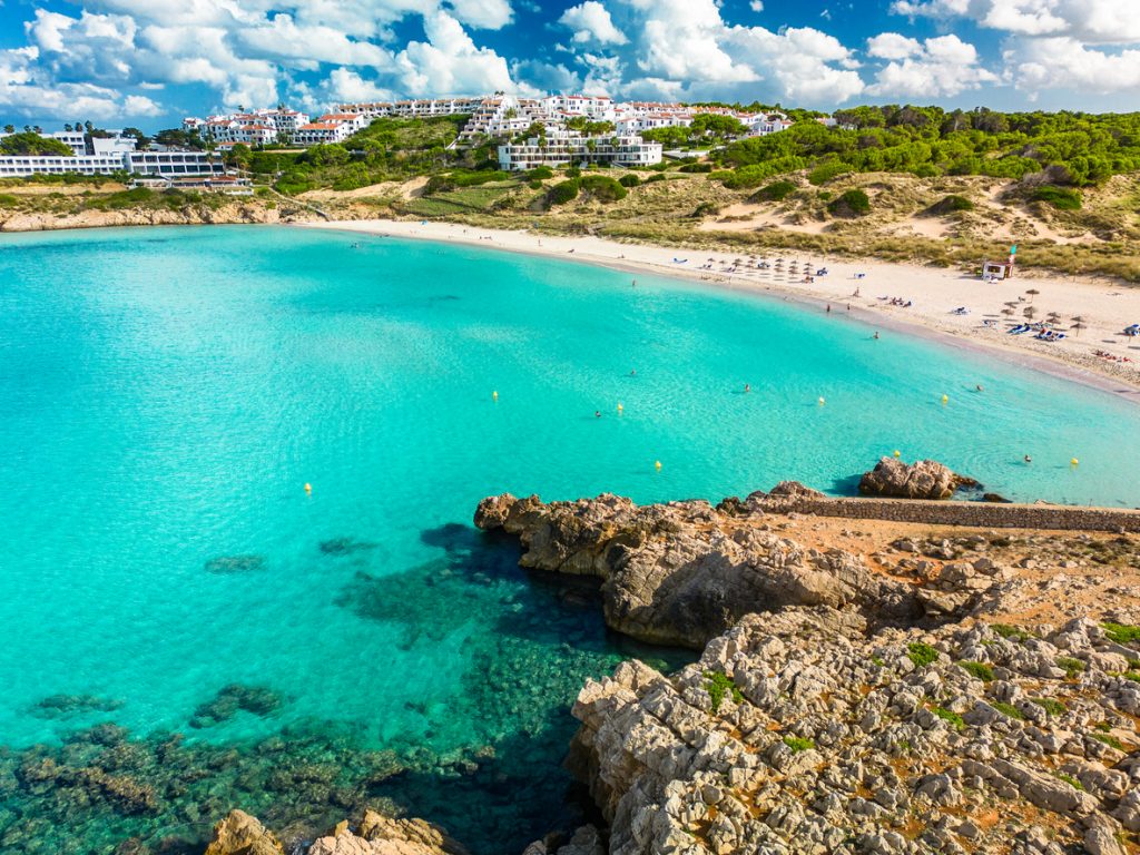 Die Bucht Son Saura zeigt einen langen, hellen Strand mit türkisfarbenem Meer und Dünen im Hintergrund.