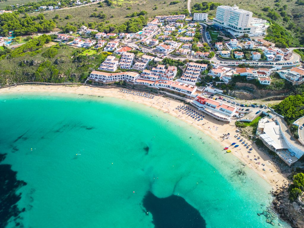Arenal d’en Castell zeigt eine breite, geschwungene Sandbucht mit klarem Wasser und Hotels am Hang.