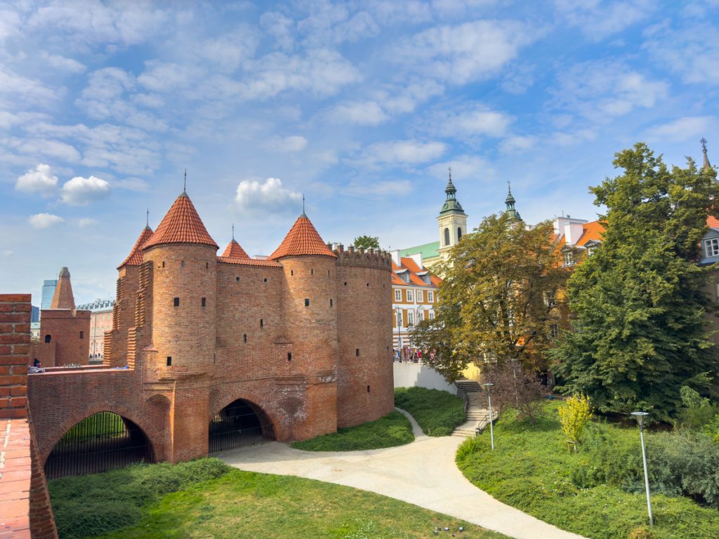 Die rote Backsteinbarbakane in Warschau zeigt Türme, Bögen und einen grünen Weg davor.