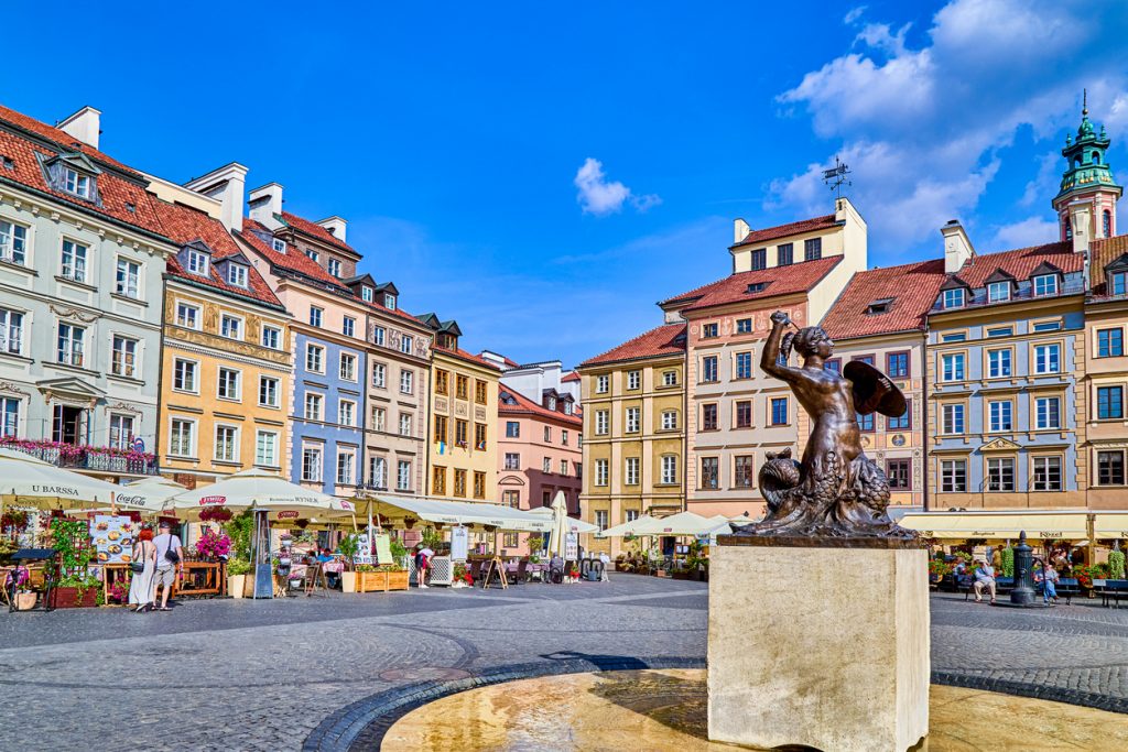 Die Skulptur der Warschauer Seejungfrau steht vor bunten Altstadthäusern und Caféterrassen.