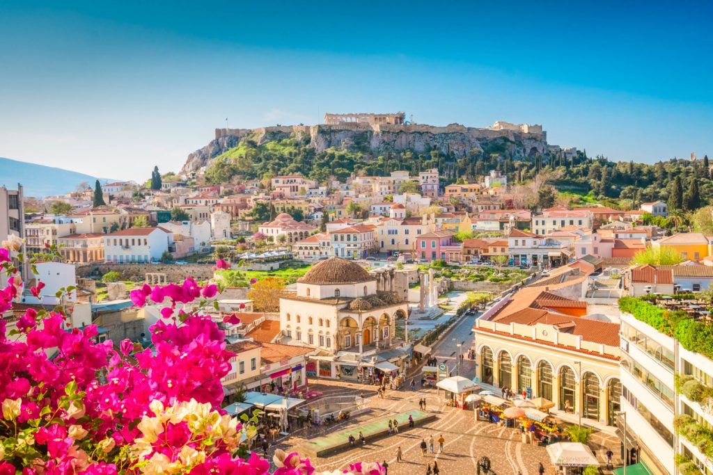 Blick über Athen mit pinker Bougainvillea im Vordergrund und der Akropolis mit dem Parthenon auf dem Hügel im Hintergrund.
