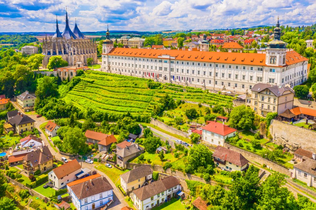 Blick über Kutná Hora mit großer gotischer Kirche, langem Klostergebäude und grünen Terrassen im Vordergrund.