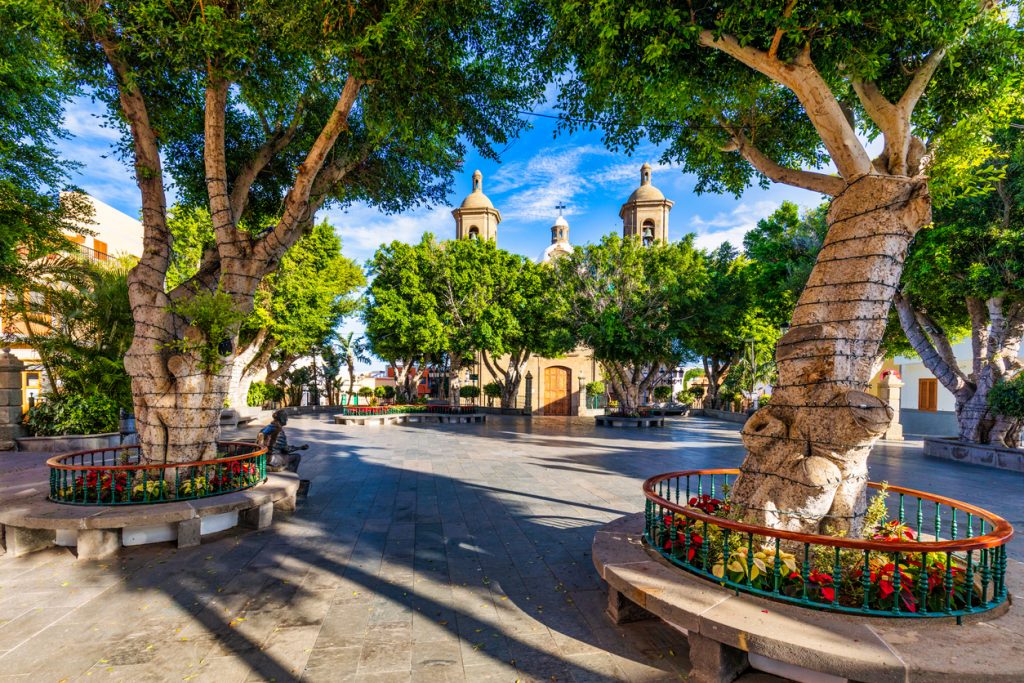 Ein ruhiger Platz liegt unter großen Bäumen mit einer Kirche dahinter.