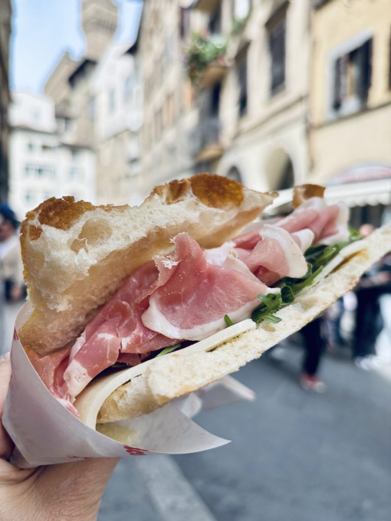 Ein belegtes Panino mit Schinken und Käse wird in der Hand vor einer Straße in Florenz gehalten.