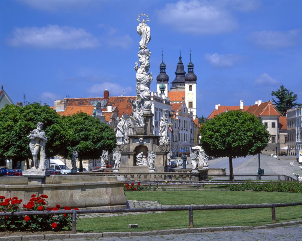 Mariensäule mit Figuren in Telč vor roten Dächern und Kirchtürmen unter blauem Himmel.