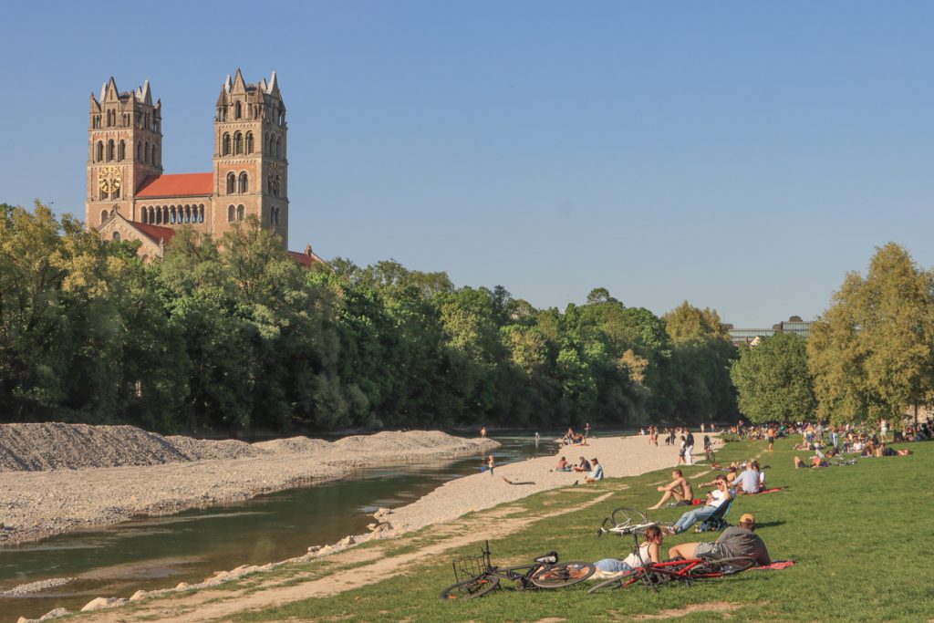 Menschen liegen an den Isarauen in München, während im Hintergrund Kirchtürme über den Bäumen auftauchen.