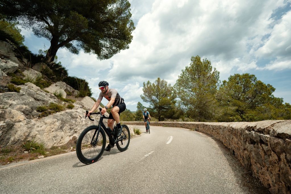 Zwei Rennradfahrer fahren auf einer schmalen Straße durch Felsen und Pinien.
